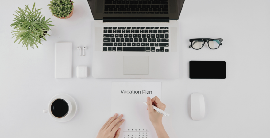 View of desk with a computer and hands writing vacation plan on a piece of paper.