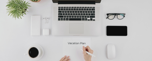 View of desk with a computer and hands writing vacation plan on a piece of paper.